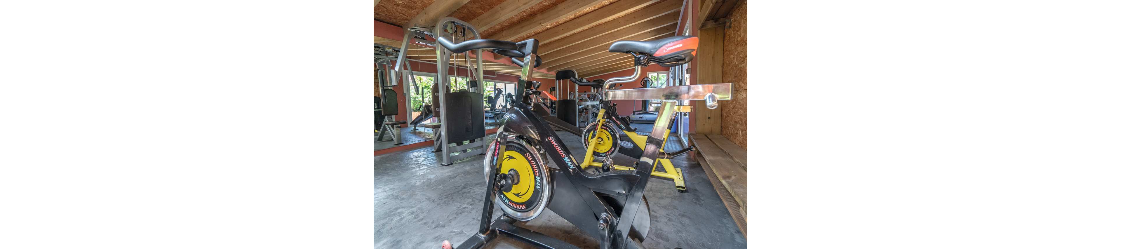 A stationary exercise bike is prominently positioned in a spacious indoor gym. Additional fitness equipment, including another bike and weight machines, can be seen in the background. The room features exposed wooden beams, large windows, and concrete flooring, creating an airy and functional workout space. Bright colors on the equipment add visual interest to the otherwise neutral interior.