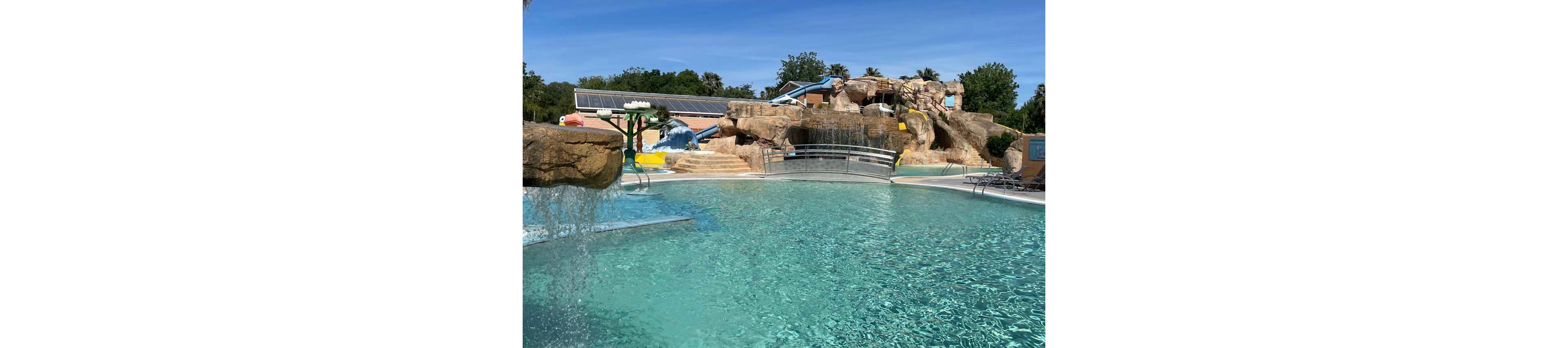 An outdoor water park pool sparkles under a clear blue sky. Rock formations with integrated water slides and tunnels rise in the background. Gentle waterfalls and fountains pour into the turquoise swimming pool in the foreground. Lounge chairs and surrounding greenery suggest a relaxed, family-friendly recreational setting.