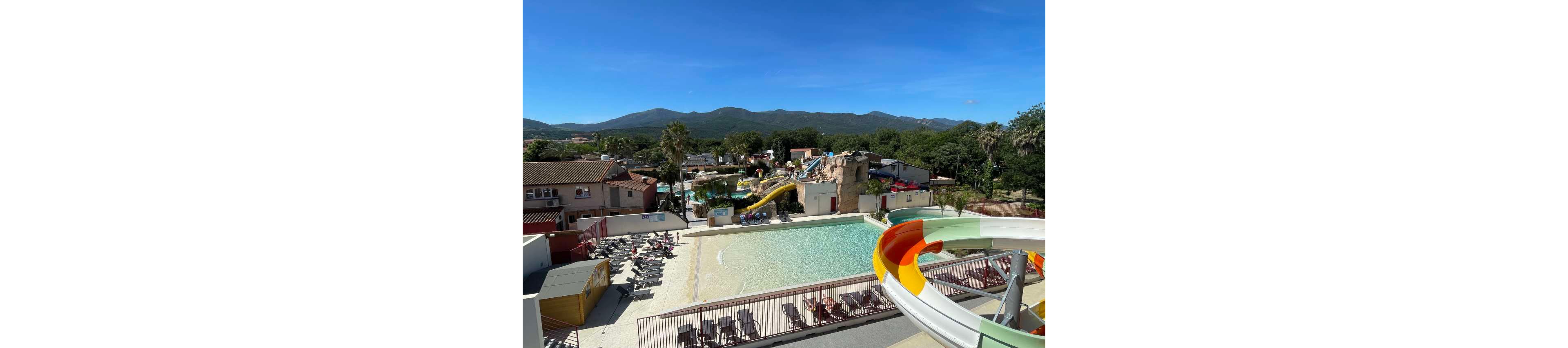 An outdoor water park features a large shallow pool surrounded by lounge chairs under a clear blue sky. Colorful water slides curve into the pool area, including a prominent red, yellow, and white slide in the foreground. Buildings, palm trees, and greenery frame the complex, with mountains visible in the distant background. The bright sunlight and clear weather create a relaxed vacation atmosphere.