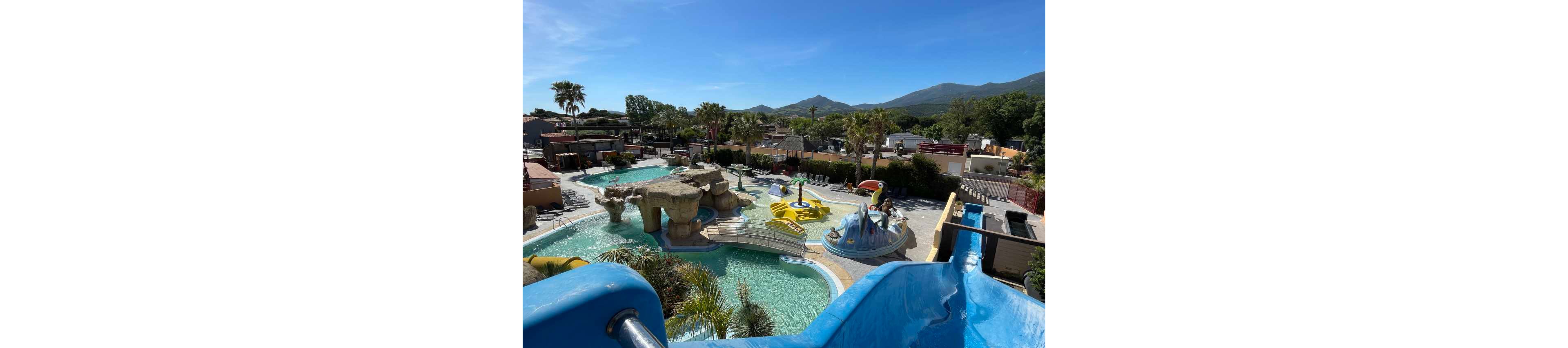 An outdoor water park features winding blue slides leading into clear turquoise pools. The scene includes rock formations, palm trees, and playful animal-shaped structures in the water. A few people enjoy the attractions under bright sunshine, with lounge chairs arranged around the pools. In the background, low buildings and distant green mountains complete the holiday resort setting.