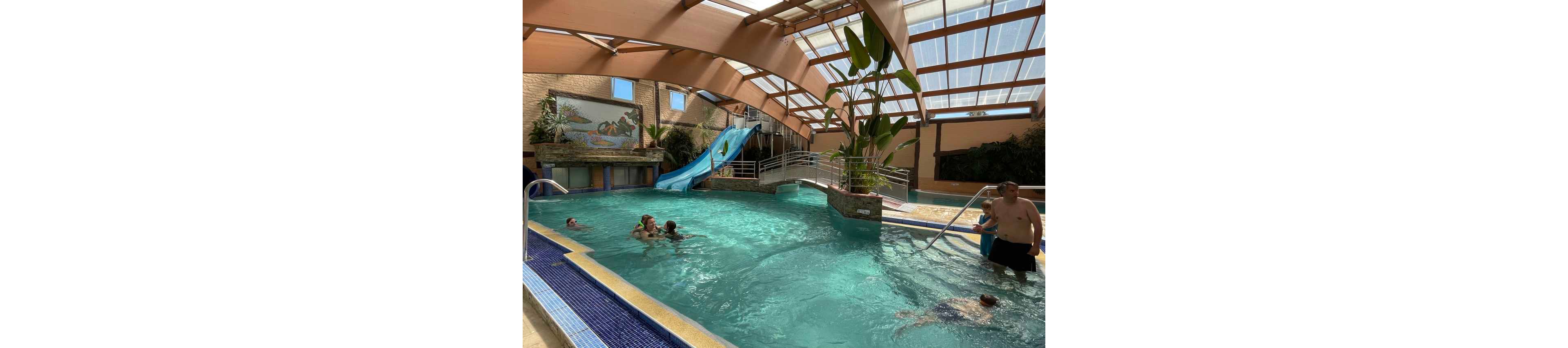 Several people swim and play in a bright indoor swimming pool. A blue waterslide curves into the pool beside a raised deck and railings. Sunlight filters through the transparent roof, illuminating the turquoise water and surrounding plants. Adults and children relax and have fun in this family-friendly aquatic setting.