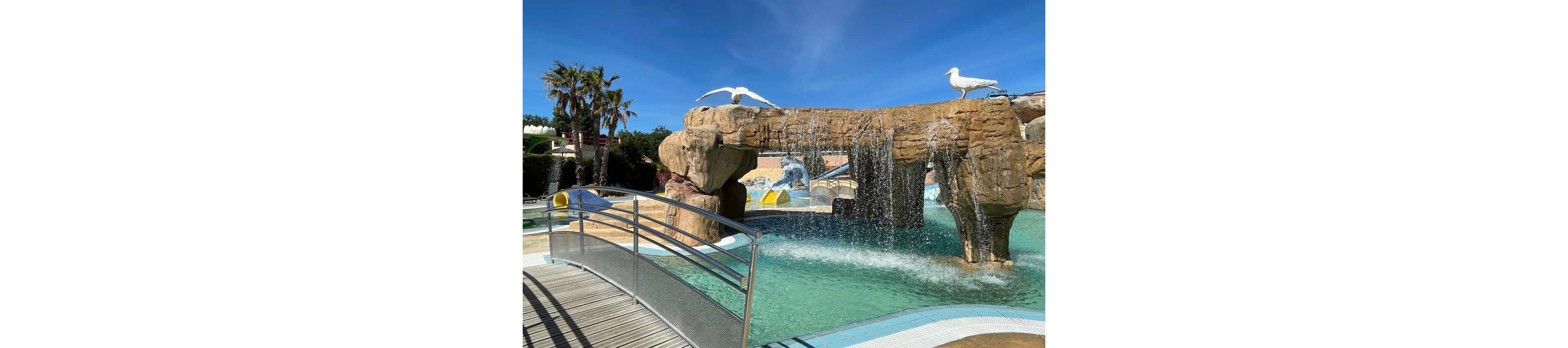 An outdoor water park features a large artificial rock structure with cascading waterfalls into a clear turquoise pool. A curved metal footbridge crosses the water in the foreground, leading toward the rocky archway. White bird statues stand on top of the rocks, and palm trees rise against a bright blue sky in the background. The scene suggests a warm, sunny day ideal for leisure and family fun.