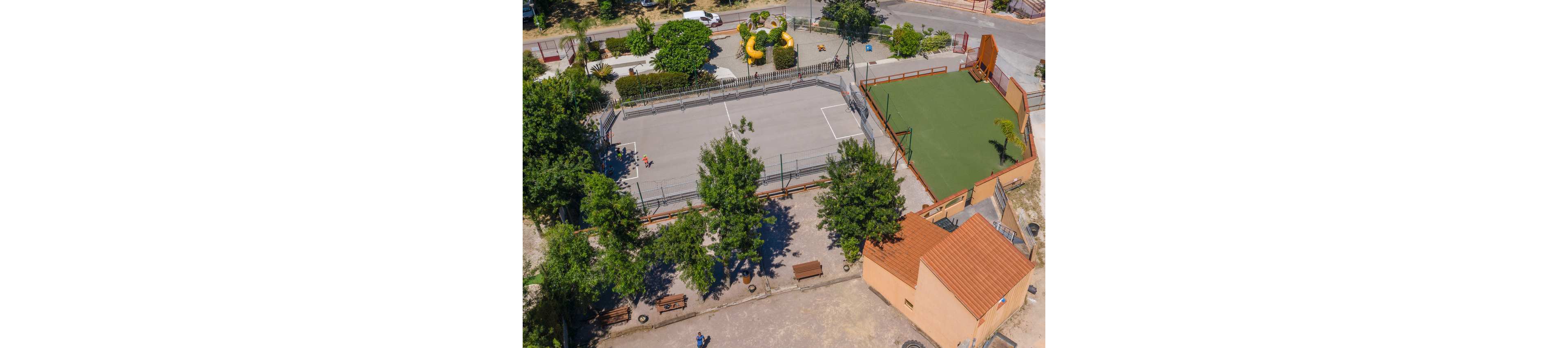 Aerial view of an outdoor recreation area featuring two adjacent sports courts. One court has a gray hard surface with painted lines, while the other has a green artificial turf field enclosed by fencing. Trees and benches surround the courts, and a small building with a tiled roof sits nearby. In the background, a playground with colorful structures and nearby streets are visible.