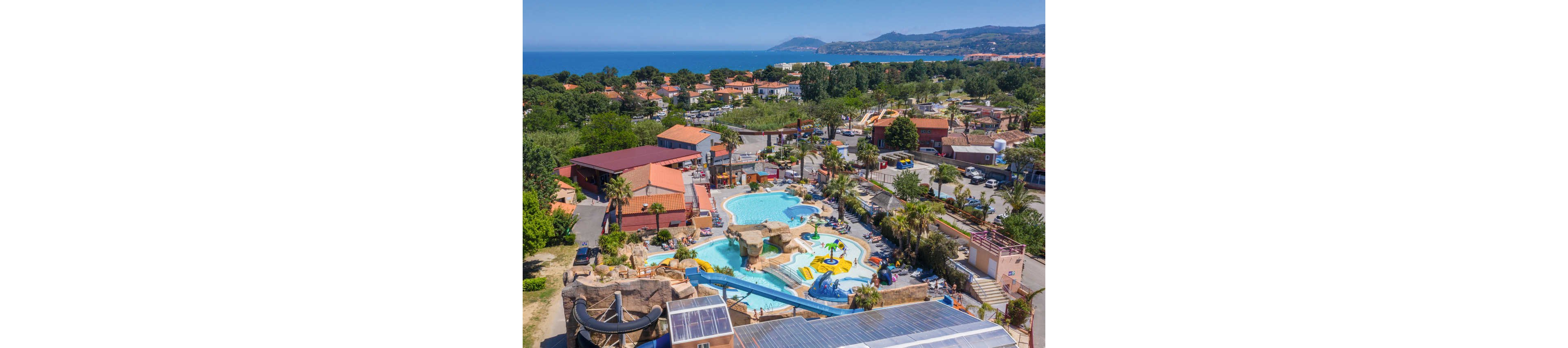 A large outdoor water park with multiple pools and slides is seen from above in a coastal resort town. The complex includes winding water slides, rock formations, and children’s play areas surrounded by palm trees and sun loungers. Red-roofed buildings and campground-style accommodations encircle the aquatic center. In the distance, the blue sea and rolling hills frame the horizon under a clear sky.