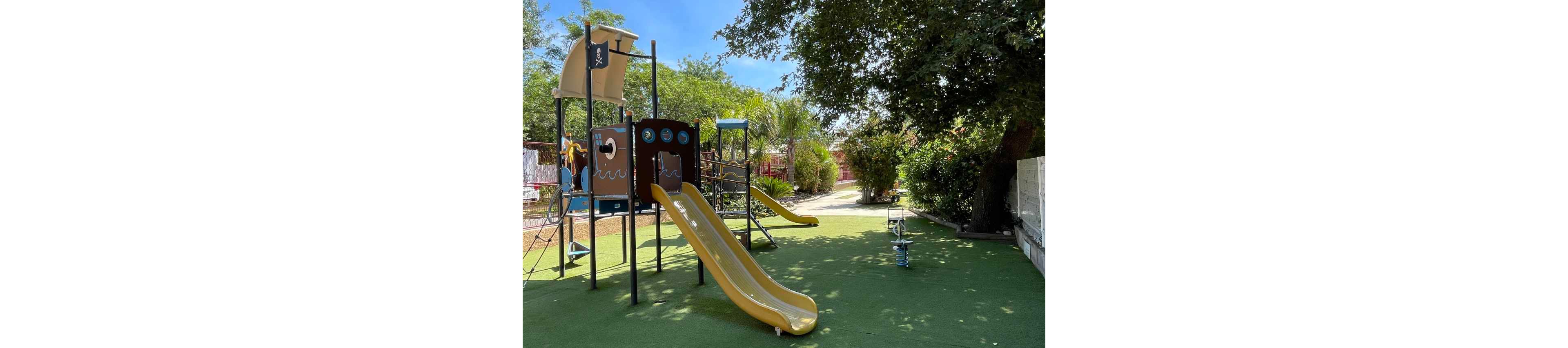 An empty playground features a pirate ship themed play structure with a bright yellow slide. The equipment stands on green artificial turf surrounded by trees and shrubs. A small spring rider is visible in the foreground, and another slide appears in the background. The scene is lit by clear, sunny weather, creating a cheerful and inviting atmosphere.