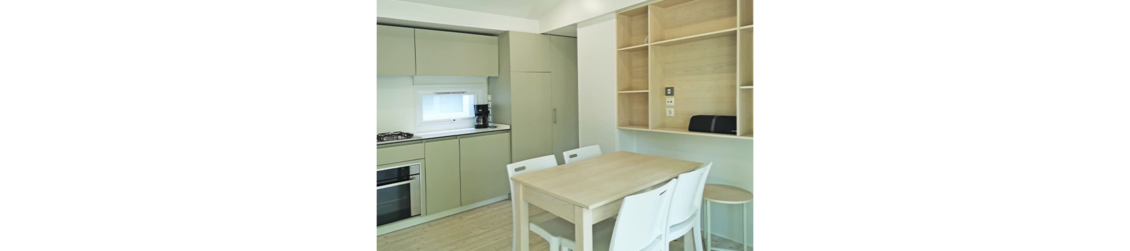 A compact modern kitchen features sleek light-colored cabinets and integrated appliances. A wooden dining table with white chairs sits in the center of the room, creating a simple and functional eating area. Open shelving and built-in storage line the wall, adding to the minimalist aesthetic. Soft natural light enters through a small window above the countertop, enhancing the clean and airy feel of the space.