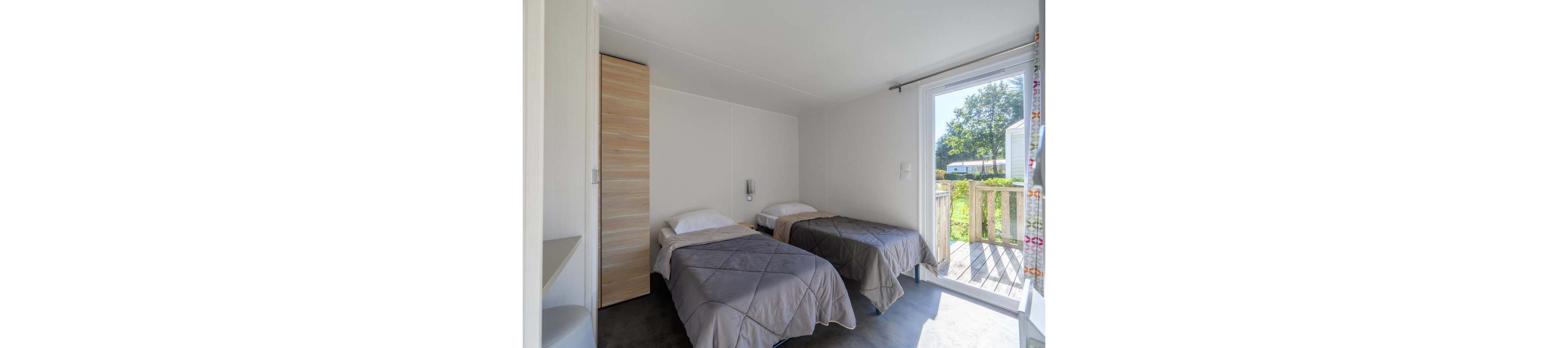 Interior view of a small bedroom featuring two single beds with gray quilts and white pillows. The room has minimalist white walls, a light wood wardrobe, and a simple bedside table between the beds. Sunlight enters through a large glass door that opens onto a wooden terrace and green outdoor area. The overall atmosphere is clean, modern, and inviting, suitable for vacation lodging or guest accommodation.