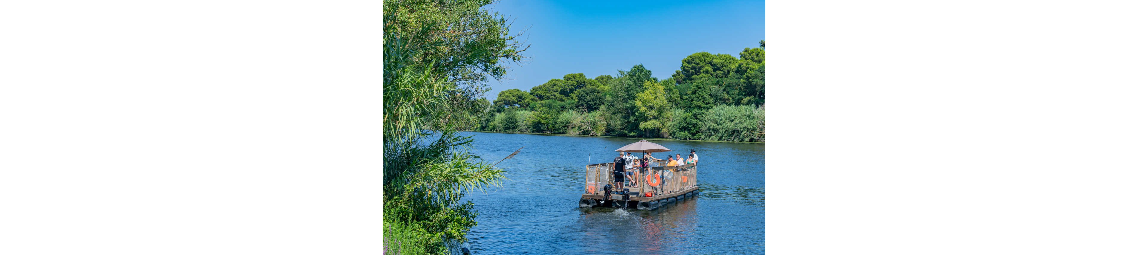 A small open ferry boat carries a group of people across a calm river. The passengers stand and sit under a sunshade while enjoying the scenic ride. Lush green trees and reeds line the riverbanks under a clear blue sky. The scene suggests a relaxed outdoor excursion in a natural setting.
