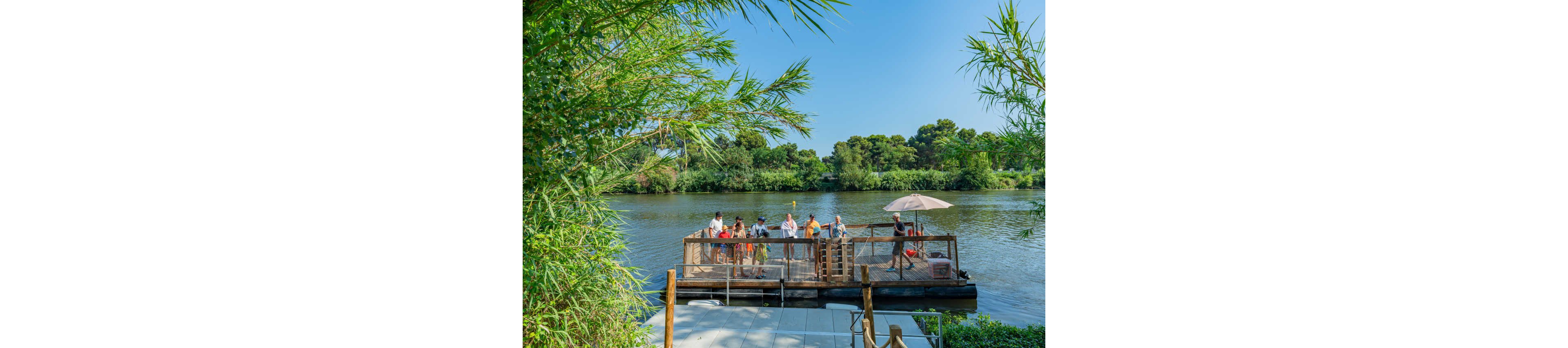 A group of people stands and sits on a wooden deck overlooking a calm river. The deck is surrounded by lush green trees and vegetation on both banks. An umbrella provides shade while visitors enjoy the sunny weather and scenic water view. The scene suggests a peaceful outdoor gathering in a natural riverside setting.