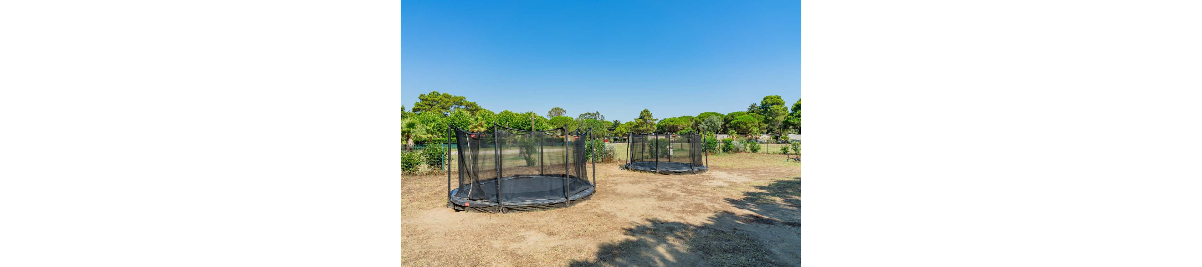 Two round ground-level trampolines with safety nets stand in an open outdoor area. The trampolines are placed on dry, bare earth surrounded by green trees and shrubs. A clear blue sky and bright sunlight create a warm, summery atmosphere. The scene suggests a recreational play zone in a park or large backyard.
