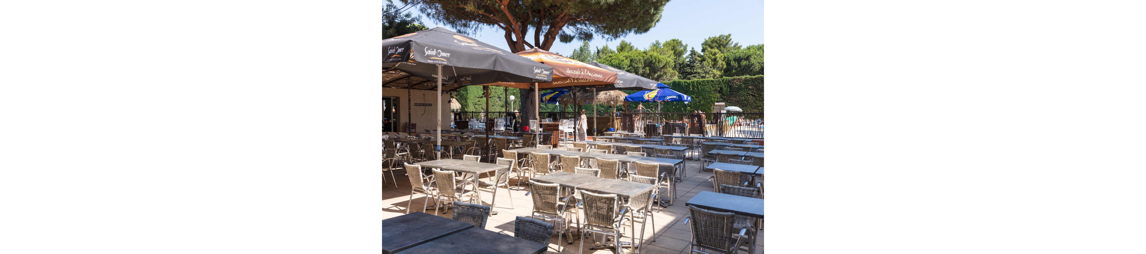 An empty outdoor café terrace is arranged with rows of metal tables and chairs under large sun umbrellas. The scene is set on a paved patio beside a fenced area, with a few people visible in the background. Tall trees and dense green hedges surround the space, creating a shaded, relaxed atmosphere. The bright daylight and clear sky suggest a warm, sunny day at a leisure or holiday destination.