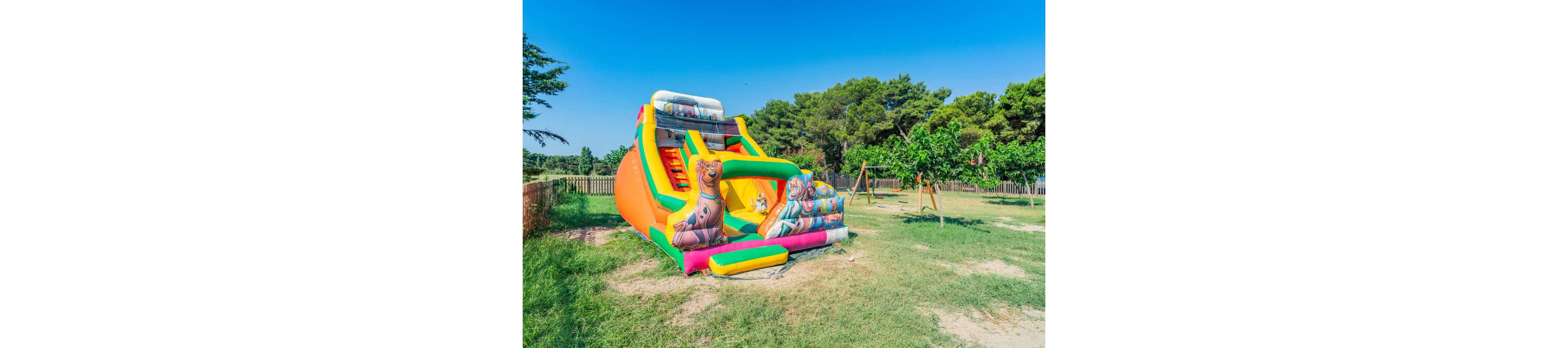 A large colorful inflatable slide stands on a grassy area in an outdoor playground. The slide features bright orange, green, and yellow sections with cartoon animal characters printed on the front. Surrounding the inflatable are trees, a wooden fence, and a swing set under a clear blue sky. The scene suggests a fun, family-friendly environment on a sunny day.