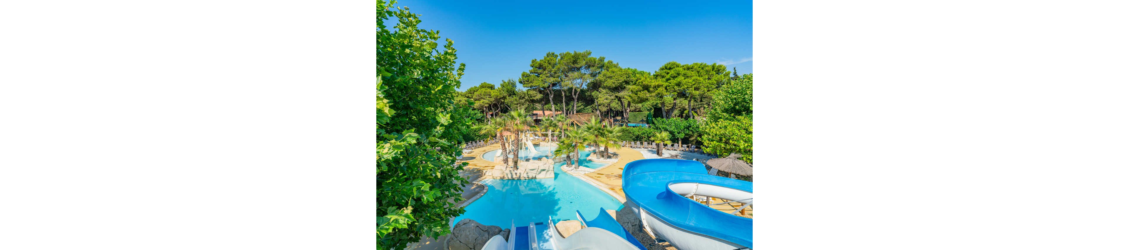An outdoor water park features winding blue water slides leading into a clear turquoise pool. The pool area is surrounded by lush green trees and palm trees, creating a tropical atmosphere. Sun loungers and umbrellas are arranged around the water, suggesting a relaxing holiday setting. The bright blue sky and vivid colors emphasize a warm, sunny summer day.