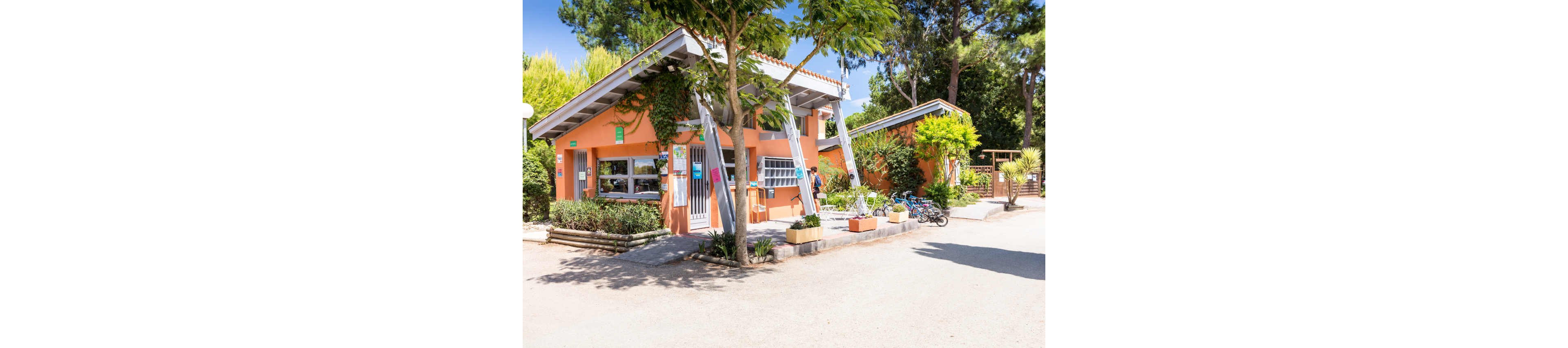 A small orange reception building stands in a sunny outdoor campsite. The structure features a sloped roof, large windows, and potted plants arranged along the entrance. Several bicycles are parked nearby, suggesting a family-friendly holiday setting. Tall trees and lush greenery surround the area, creating a relaxed summer atmosphere.