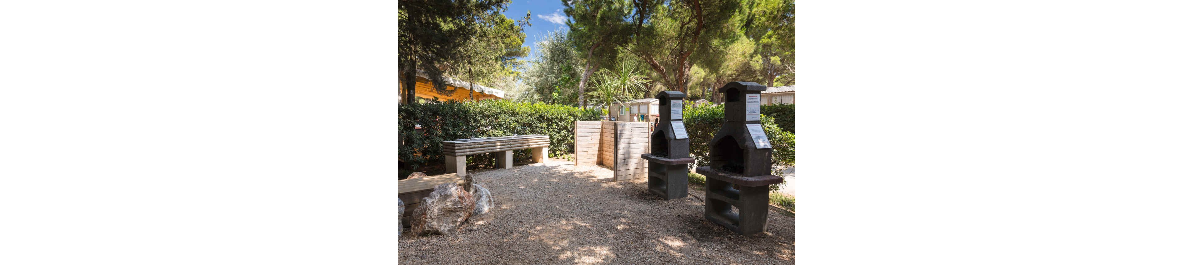 Image of an outdoor barbecue area featuring two stone or concrete grills. The setting appears to be a campsite or holiday park surrounded by dense green hedges and tall trees. A wooden privacy screen and a long bench provide functional elements within the gravel-covered ground. Sunlight filters through the foliage, creating a relaxed and inviting atmosphere for outdoor cooking.