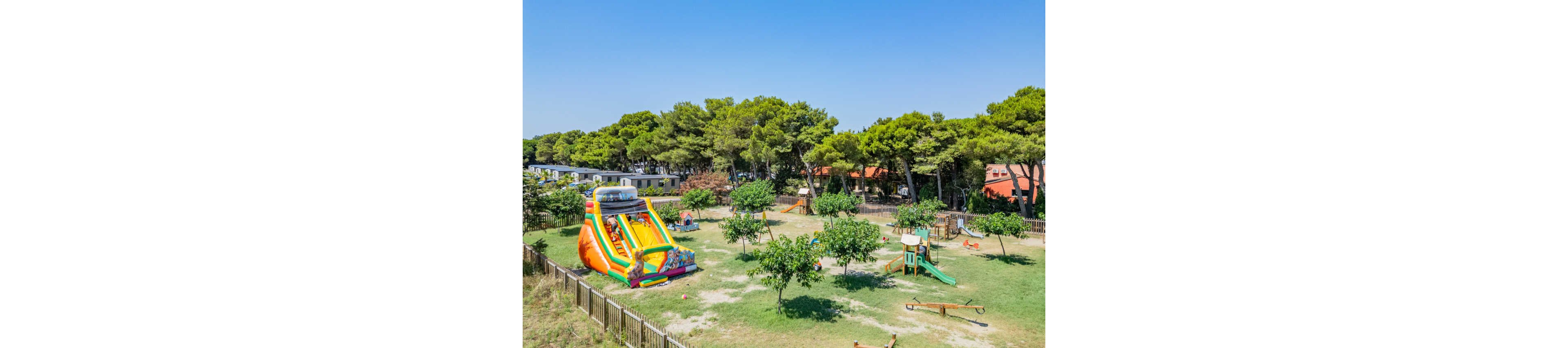 An expansive outdoor playground features a large colorful inflatable slide as its main attraction. The area is set in a grassy field surrounded by trees and nearby buildings under a clear blue sky. Additional play structures such as swings, slides, and climbing frames are scattered around the fenced space. The scene suggests a family-friendly recreational area in a warm, sunny climate.