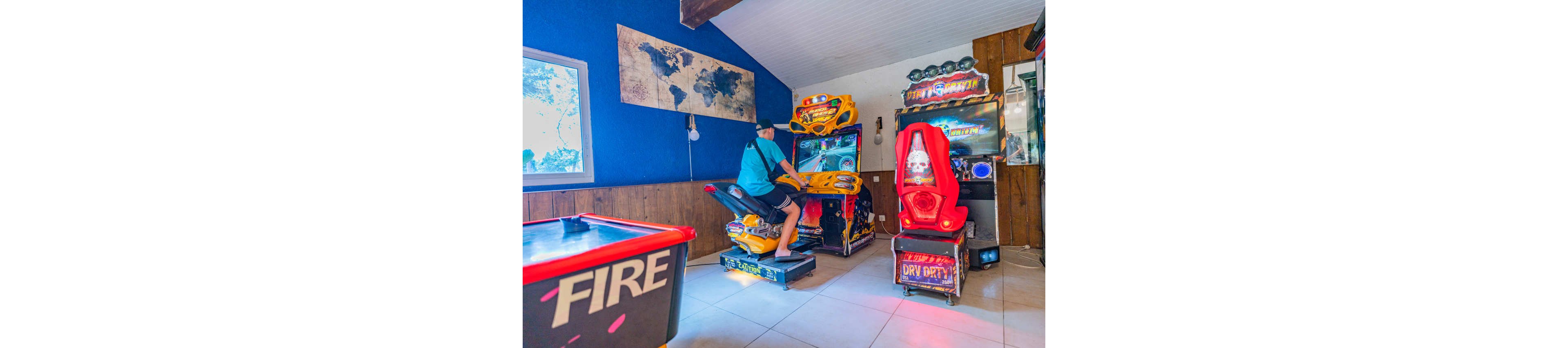 A teenage boy plays a motorcycle racing arcade game in a brightly lit game room. He sits on a yellow motorbike simulator while facing a large screen with colorful graphics. Other arcade machines, including a red driving game labeled "DRY DRFT" and an air hockey table with the word "FIRE," surround him. The room features blue walls, a world map wall art, and a relaxed, fun atmosphere.