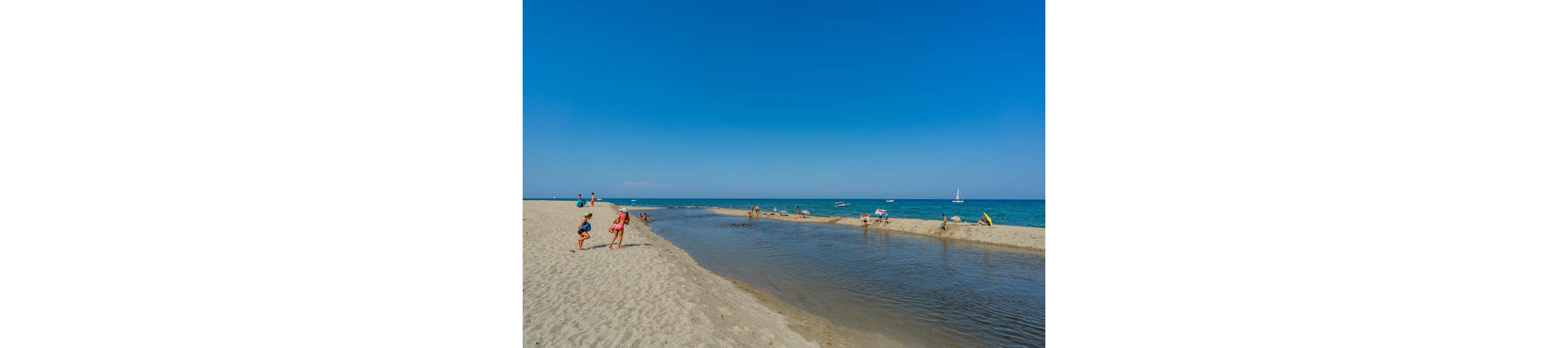 A sandy beach with a shallow inlet of water stretches toward a calm blue sea under a clear sky. Several people, including children and adults, play near the water’s edge and walk along the sand. Small boats and a distant sailboat are visible on the horizon, emphasizing a relaxed coastal setting. The bright sunlight and vivid blue tones create a warm, summery atmosphere.