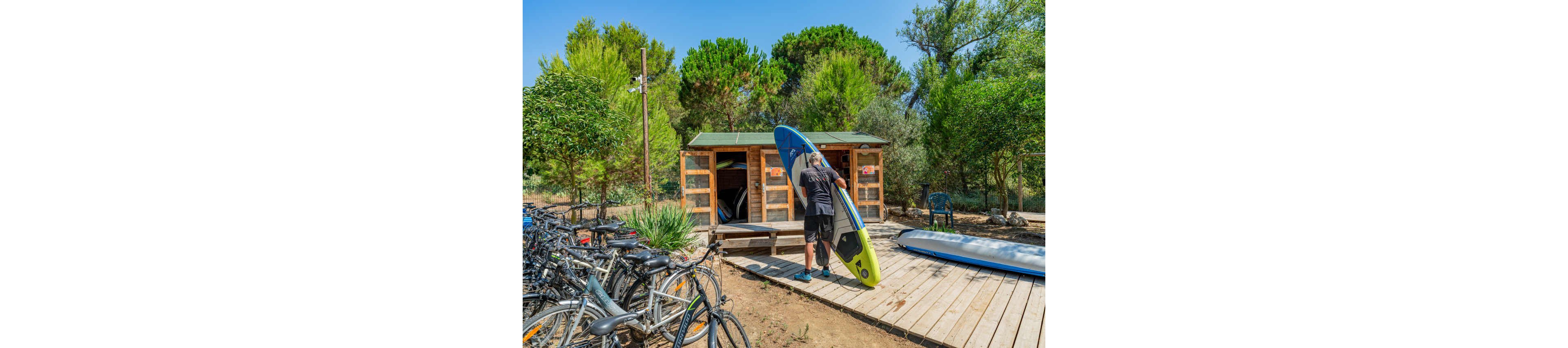 A man stands on a wooden deck lifting a paddleboard in front of a small rental hut. Several bicycles are parked nearby, suggesting multiple outdoor activity options. The scene is surrounded by lush green trees and vegetation under a bright, clear sky. The overall atmosphere feels relaxed and summery, emphasizing recreation and nature.