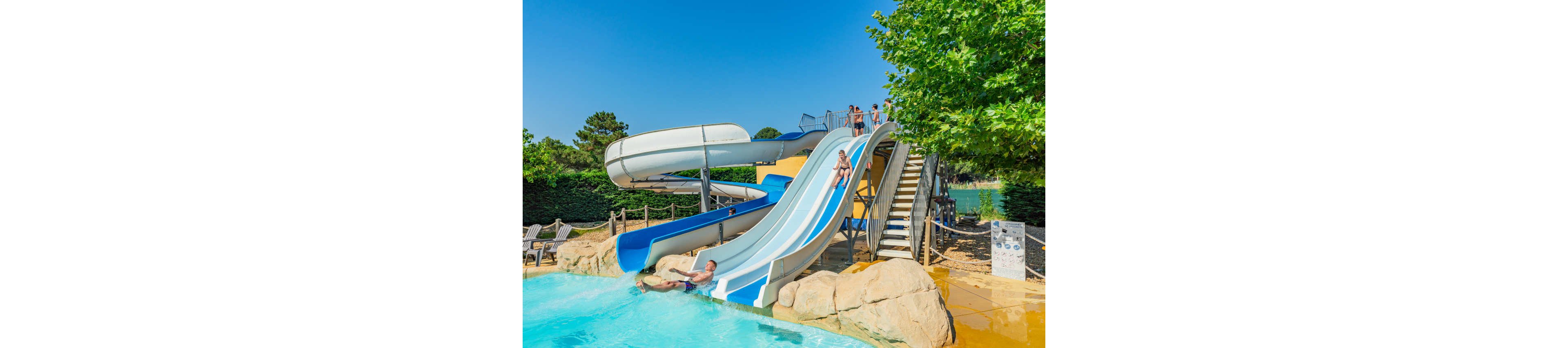 Children and adults slide down multiple water slides into a bright blue pool at an outdoor waterpark. The scene shows a curved enclosed slide and open twin slides built over artificial rocks. People climb a staircase to the top platform while others splash into the shallow water below. Lush green trees and hedges surround the attraction under a clear blue sky, creating a sunny summer atmosphere.