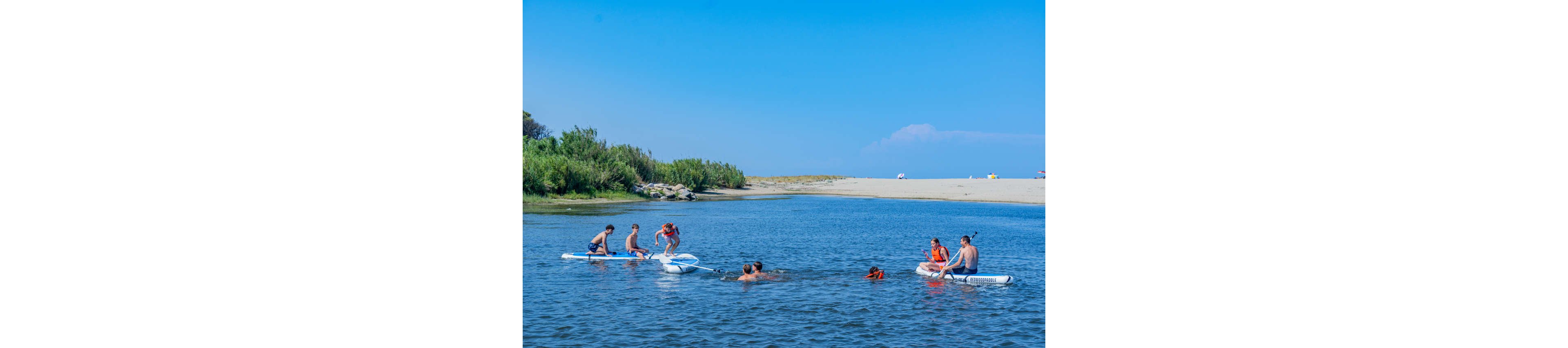 A group of children enjoy a sunny day on a calm body of water, using paddleboards and swimming near the shore. The scene is set in a shallow lagoon or river mouth with sandy banks and green reeds in the background. Clear blue sky and bright daylight create a vibrant summer atmosphere. The image conveys outdoor recreation, friendship, and playful water activities.