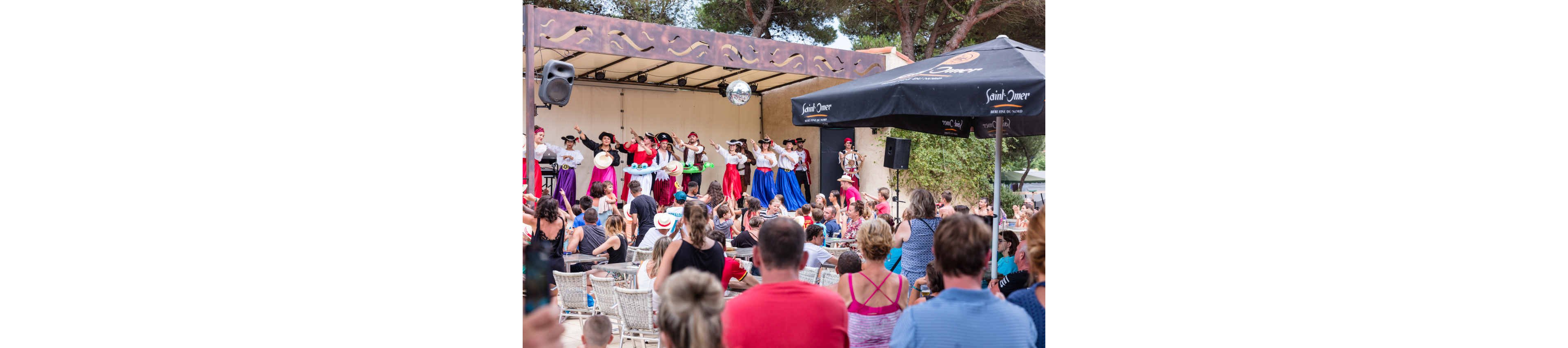 A group of dancers in colorful traditional costumes performs on an outdoor stage. The audience sits closely packed on chairs and benches, watching the show under large umbrellas. Bright skirts in red, blue, and purple contrast with white blouses and dark hats. Trees and a clear sky frame the festive summer atmosphere of the event.
