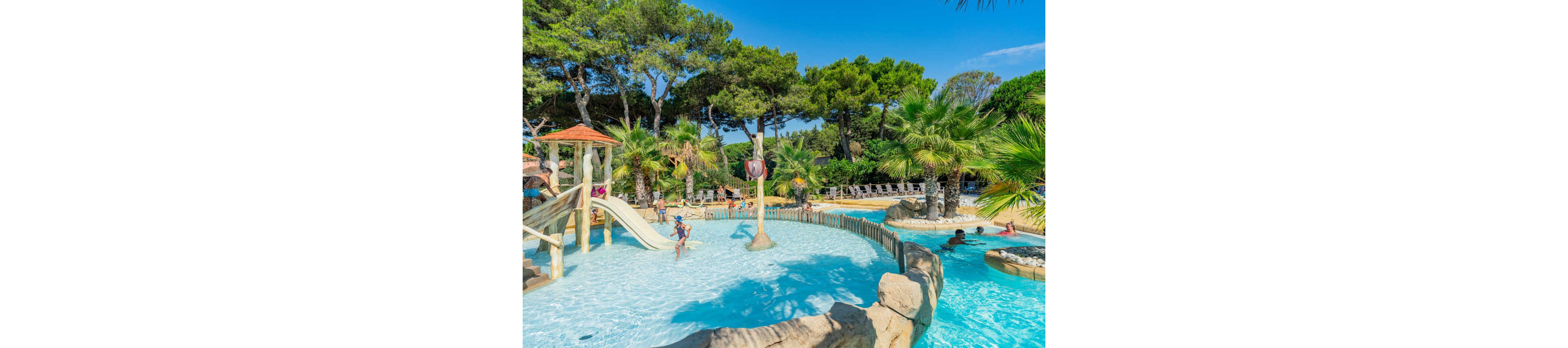 Children play in a shallow outdoor pool at a resort-style waterpark surrounded by lush trees and palm trees. A small slide and water play structures create a fun, family-friendly environment. Adults and kids relax and swim in the clear blue water under a bright, sunny sky. Lounge chairs line the background, suggesting a holiday or vacation setting.