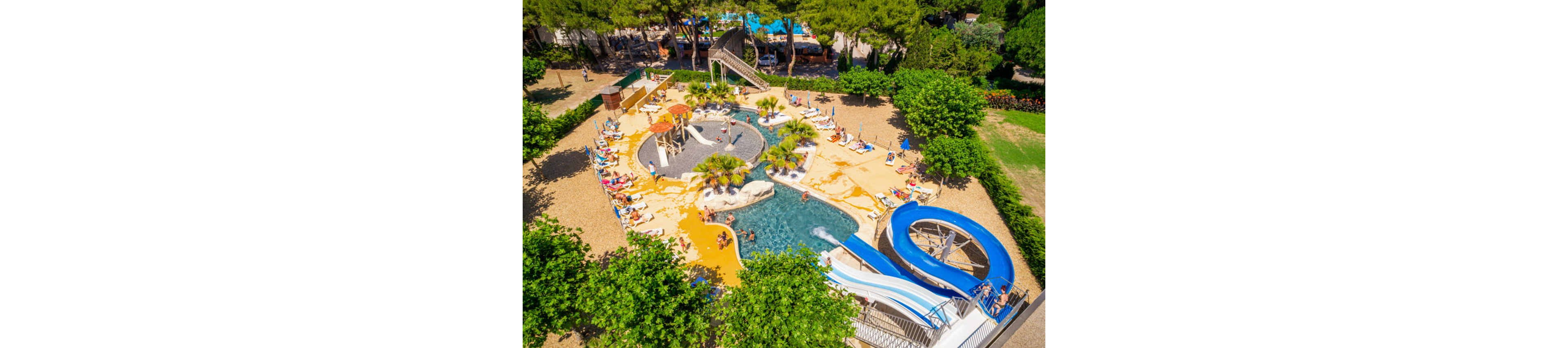 A vibrant outdoor water park is captured from above, showing pools, slides, and sunbathing areas surrounded by greenery. Families and children relax on lounge chairs and enjoy the various water attractions under bright sunlight. A large blue spiral waterslide and smaller slides lead into shallow pools with palm trees and rock features. The scene conveys a fun, summery vacation atmosphere in a recreational resort setting.
