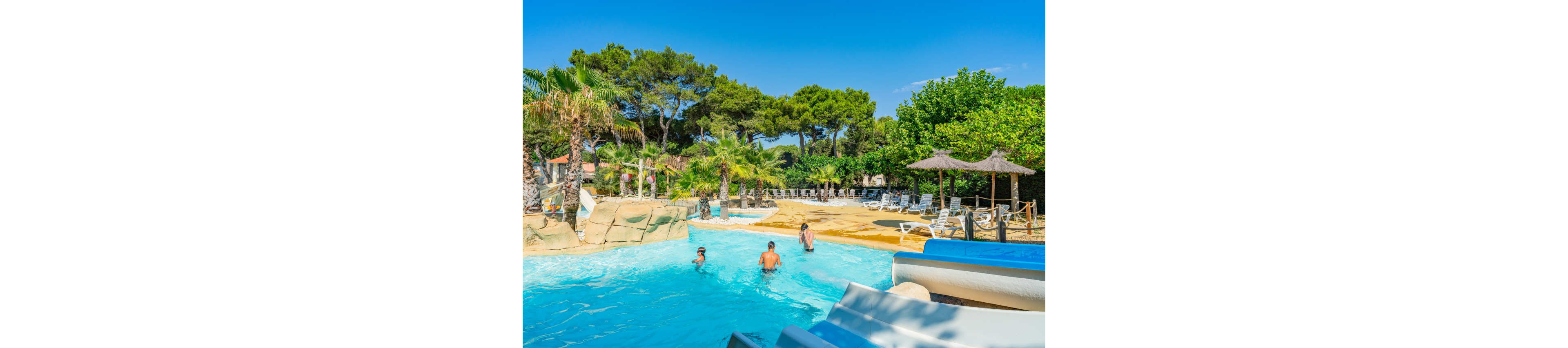 An outdoor resort swimming pool is surrounded by palm trees and lush greenery under a clear blue sky. Several people are in the water near the base of a waterslide, enjoying a sunny day. In the background, numerous lounge chairs and straw parasols line the poolside deck. The scene evokes a relaxed vacation atmosphere in a warm, tropical-style setting.