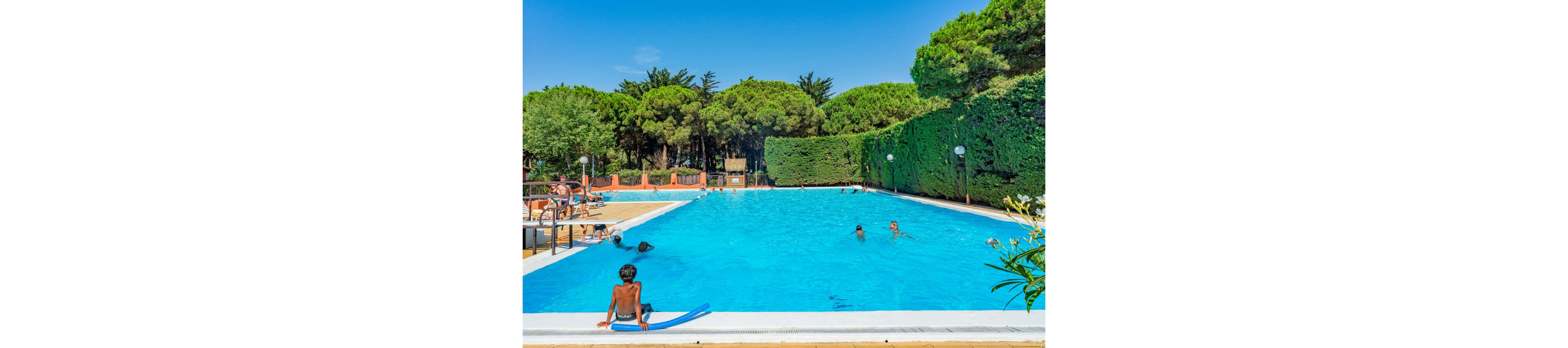 A large outdoor swimming pool is surrounded by lush green trees and hedges under a clear blue sky. Several people are swimming and playing in the bright blue water, while others relax on lounge chairs along the poolside. A child sits at the pool edge with a foam pool noodle, watching the swimmers. The scene suggests a warm, sunny day at a resort or holiday park.