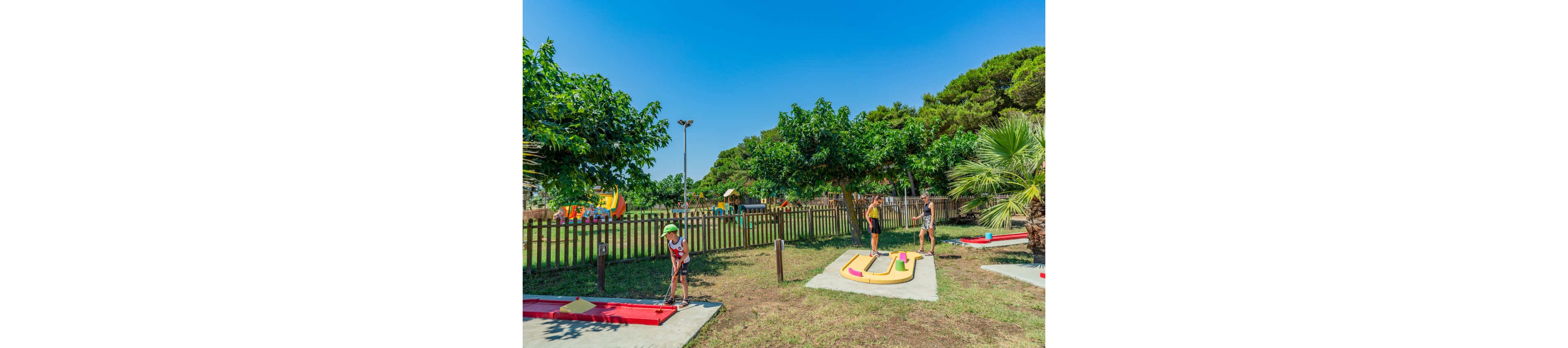 A group of people enjoy a game of mini golf on a bright, sunny day. The scene is set in an outdoor recreation area with lush green trees, grass, and a wooden fence. Colorful mini golf obstacles and putting lanes stand out against the natural surroundings. In the background, additional play structures suggest a family-friendly park environment.