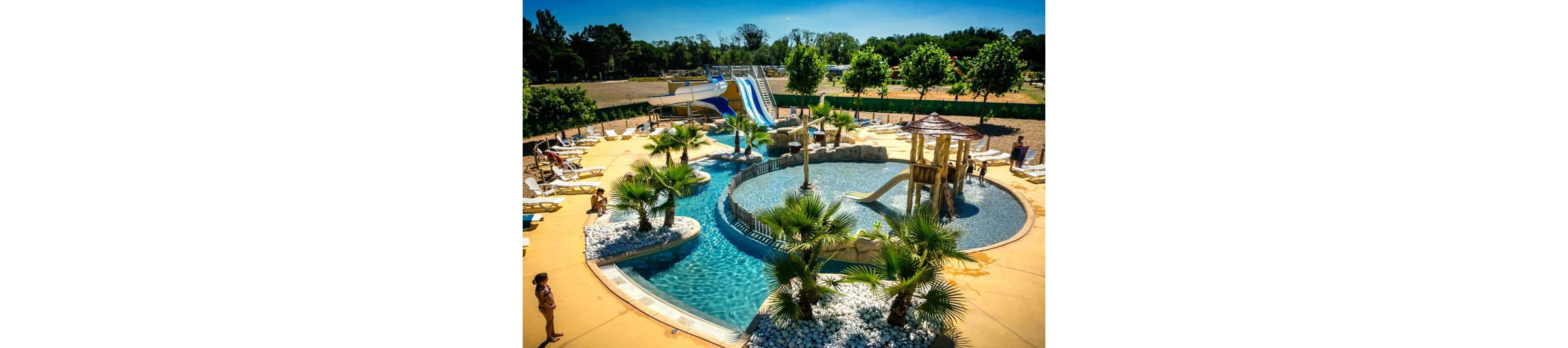 An outdoor water park features winding pools, palm trees, and a central shallow play area. Several people relax on lounge chairs while others walk along the poolside under bright sunshine. Large water slides and a wooden play structure with small slides rise in the background. The scene suggests a warm, tropical-style resort focused on family recreation and leisure.