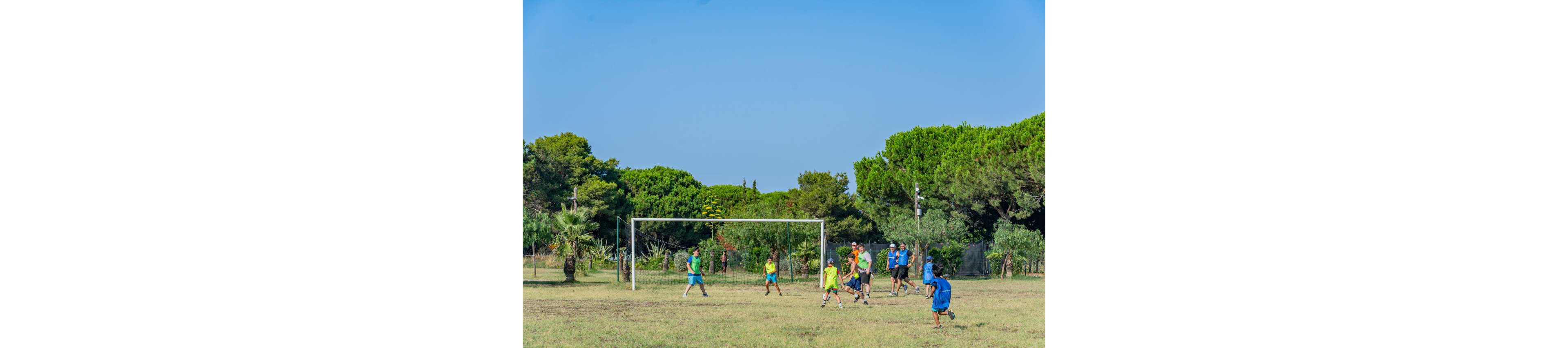 A group of children play a casual soccer game on a grassy field. They run around a simple metal goalpost under a clear blue sky. Adults or older youths stand nearby, possibly supervising or joining the game. Lush green trees and palms surround the open space, creating a relaxed outdoor park atmosphere.