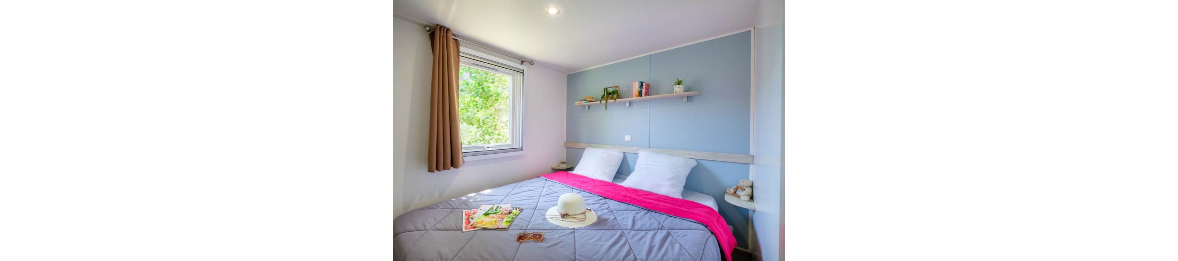 A neatly made double bed with white pillows and a gray quilted duvet dominates a compact bedroom. A bright pink throw adds a pop of color, while a white hat, sunglasses, and magazines rest on the bed. A window with beige curtains lets in natural light and greenery from outside. A floating shelf above the bed holds books and small decorative plants, creating a relaxed and inviting atmosphere.