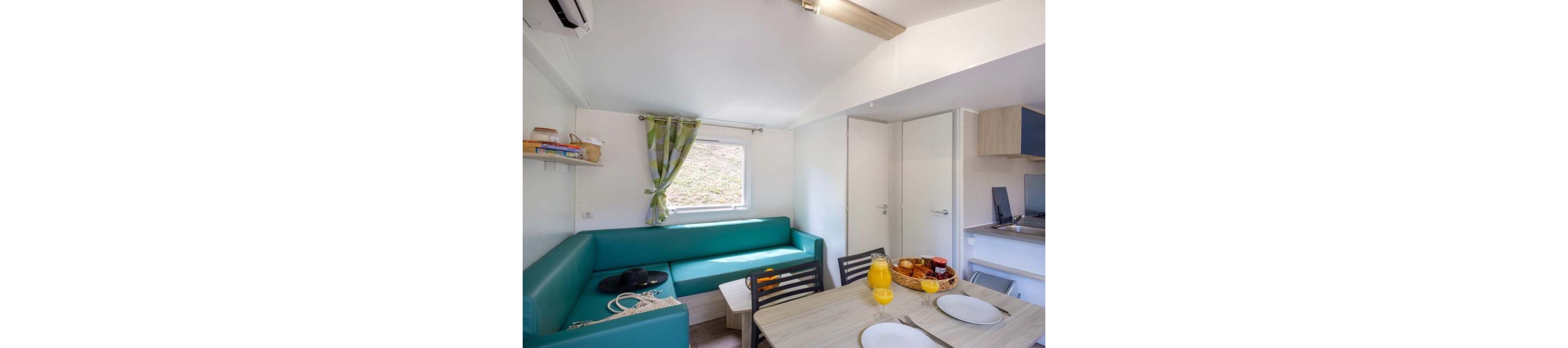 A compact living and dining area features a turquoise corner sofa beside a window with green patterned curtains. In the foreground, a light wooden table is set with white plates, cutlery, and a glass jug of orange juice. A basket of bread and pastries sits on the table, suggesting a relaxed meal. The space extends into a small kitchenette with modern appliances, creating a cozy vacation home atmosphere.