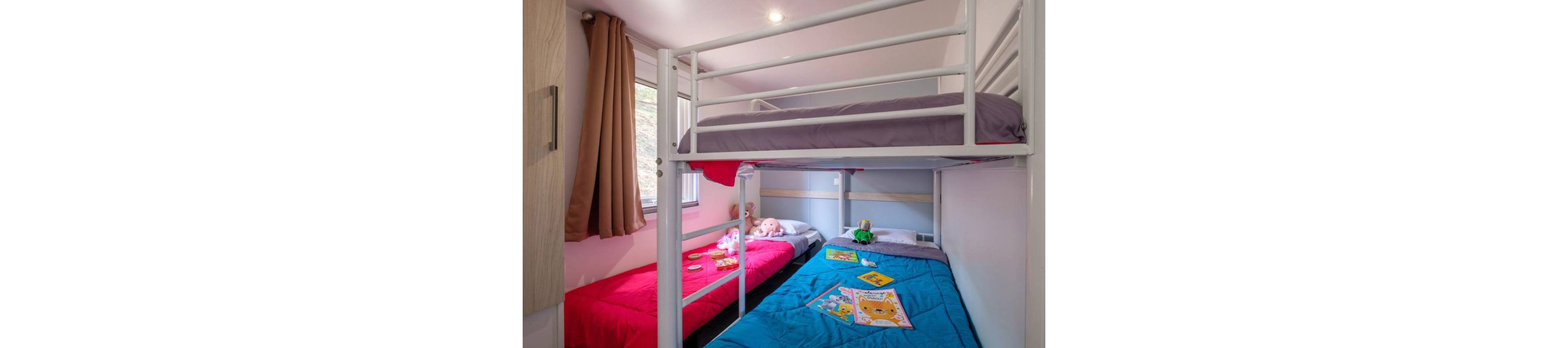 A small bedroom features a white metal bunk bed with bright bedding. The lower beds are dressed in red and blue duvets, while the top bunk has a purple cover. Children's toys and picture books are scattered on the beds, suggesting a playful and cozy space. Natural light enters through a window with brown curtains, illuminating the compact, family-friendly room.