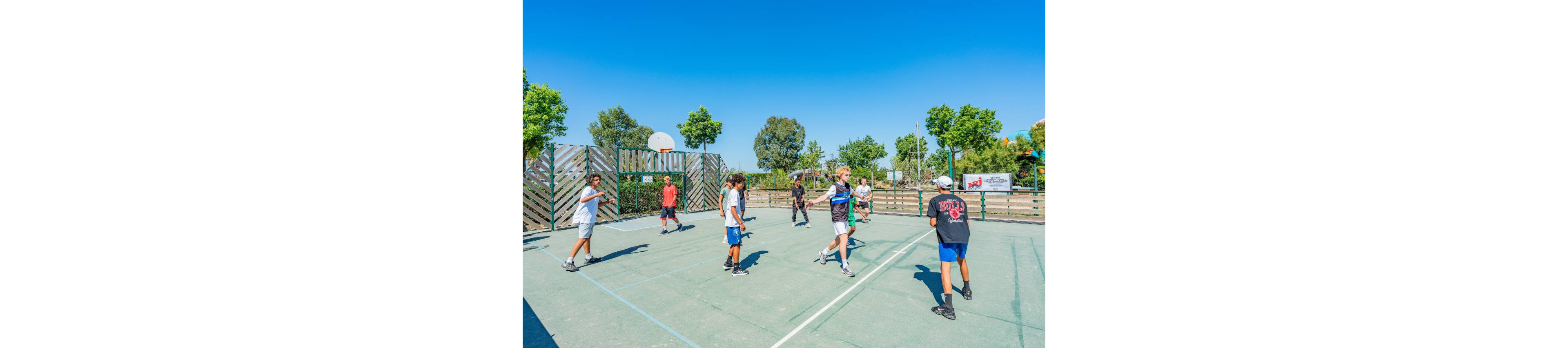 A group of teenagers play a casual game of basketball on an outdoor court. The scene is set on a sunny day with a clear blue sky and trees surrounding the court. Some players wear athletic shorts and T-shirts while others stand ready to receive the ball. A basketball hoop and fencing frame the play area, with additional sports equipment and signs visible in the background. The overall atmosphere is energetic and recreational.