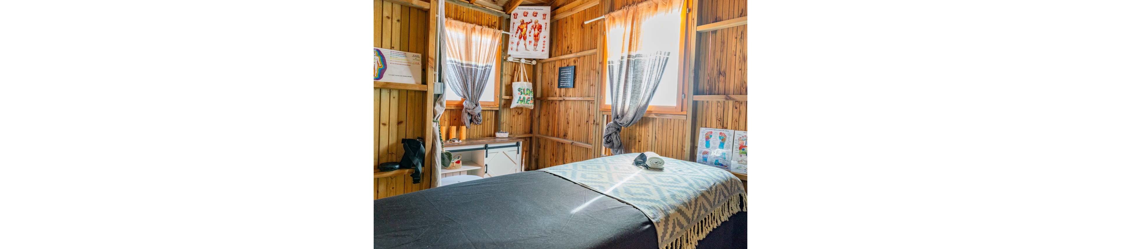 A cozy massage therapy room is set inside a wooden cabin with warm natural light streaming through curtained windows. A massage table covered with a patterned blanket and dark sheet occupies the center of the space, with a folded towel and device placed on top. Anatomical charts and posters hang on the walls, suggesting a focus on bodywork or physiotherapy. A small desk and storage area in the corner hold additional supplies, creating a functional yet relaxing environment.