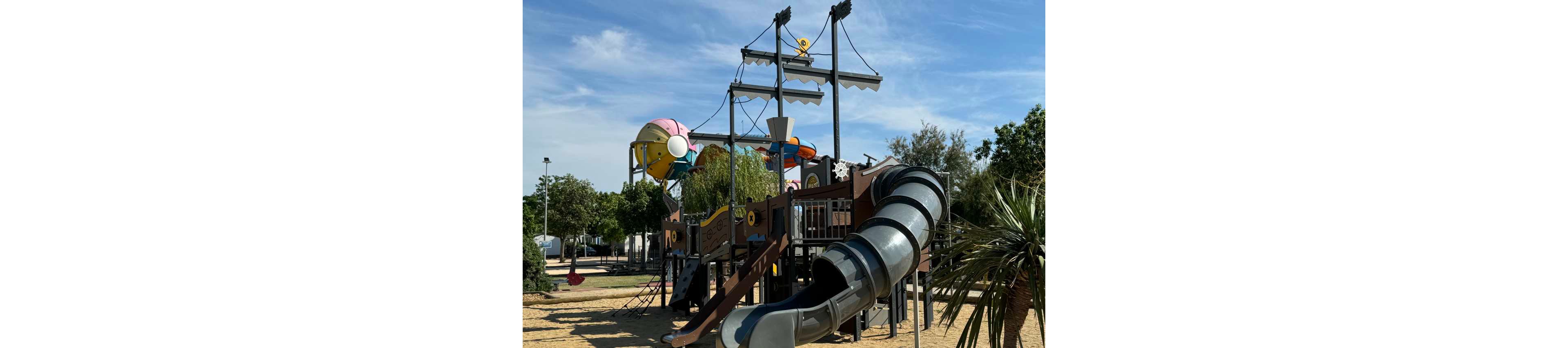 A large pirate ship themed playground structure stands in an outdoor park. The play area features multiple decks, climbing elements, and a long enclosed tube slide in the foreground. Colorful nautical details and shapes decorate the structure, with trees and open space surrounding the playground. The scene is captured on a bright, sunny day under a clear blue sky.