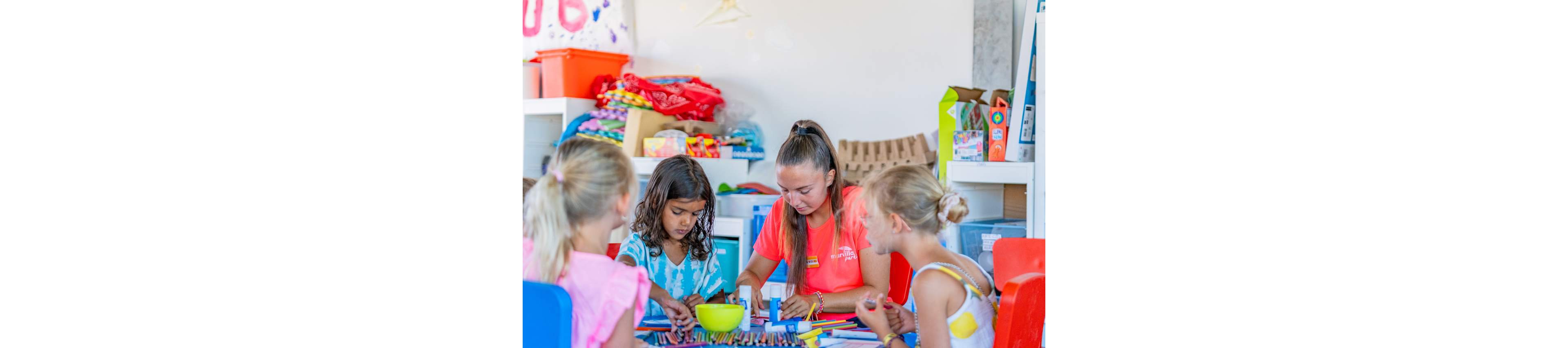 A group of children sit around a colorful table working on arts and crafts with a teacher. The setting appears to be a bright classroom or activity room filled with storage bins and supplies. Crayons, markers, and paper are spread across the table as the kids focus on their creative projects. The atmosphere is playful and educational, suggesting a summer camp or school art session.