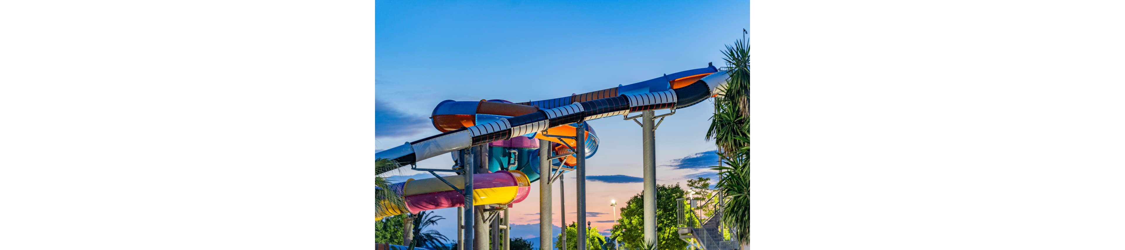 A series of large, colorful water slides curves high above the ground in an outdoor water park. The structure is supported by tall metal pillars and surrounded by tropical trees and greenery. The sky is transitioning from day to night, with soft blue and pink hues creating a serene backdrop. The scene suggests a fun, family-friendly attraction that is quiet during the evening hours.