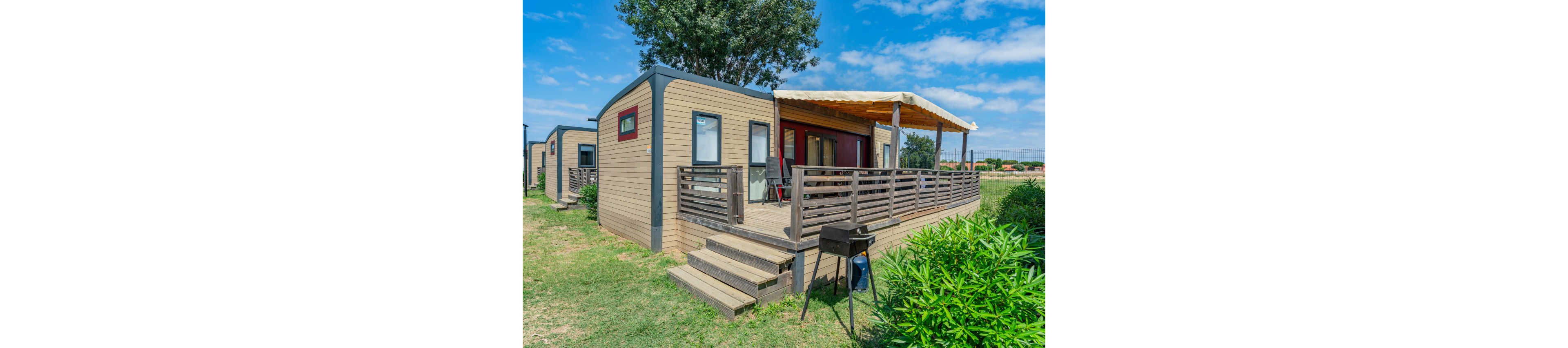 A contemporary mobile home with a wooden exterior stands in a grassy open area. The unit features a raised deck with railings, outdoor seating, and a canopy providing shade. A small charcoal grill is placed in front of the steps, suggesting outdoor cooking and leisure. Other similar cabins and a clear blue sky with scattered clouds complete the relaxed holiday setting.