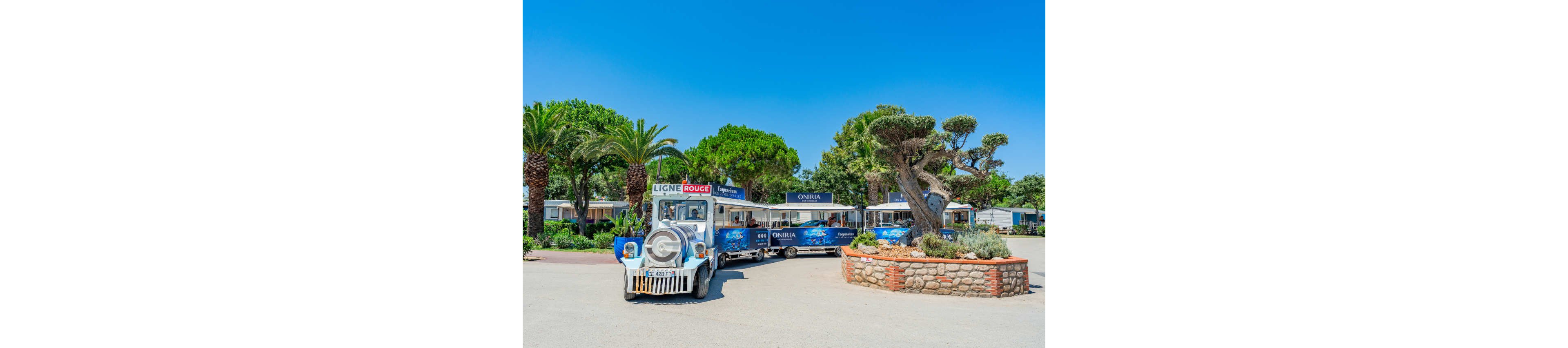 A small tourist road train painted in blue and white stands in a sunny holiday resort. The train has the text "LIGNE ROUGE" on the front and "ONIRIA" on the carriages, suggesting a themed route. It is parked on a wide paved area surrounded by palm trees, trimmed pines, and low buildings. The bright blue sky and lush greenery create a relaxed, summery atmosphere.
