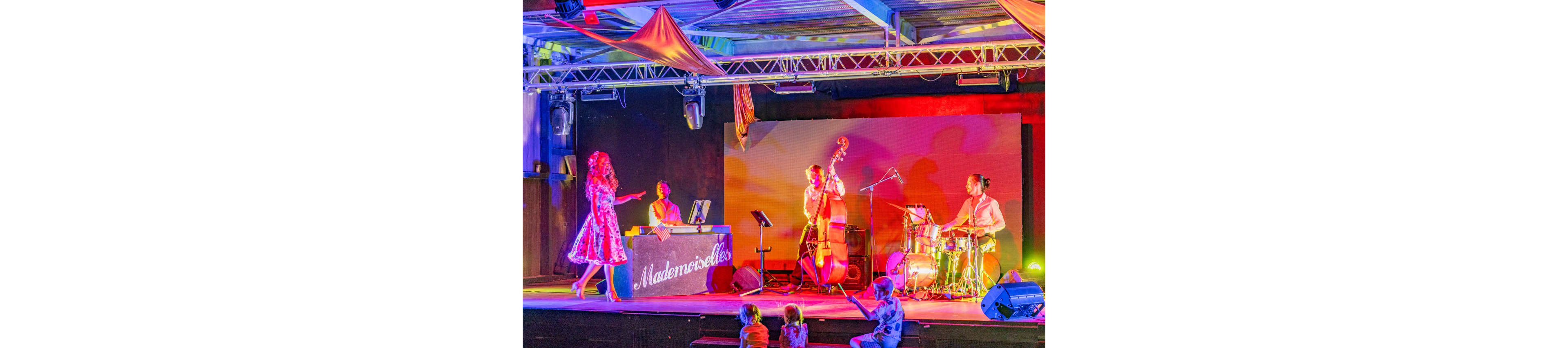 A vibrant jazz band performs on a brightly lit stage, featuring a singer, pianist, double bassist, and drummer. The scene is bathed in colorful lighting with strong yellow, pink, and blue tones. Several young children sit and watch from the floor in front of the stage, adding a family-friendly atmosphere. The word "Mademoiselles" is clearly visible on the front of the pianist's stand.