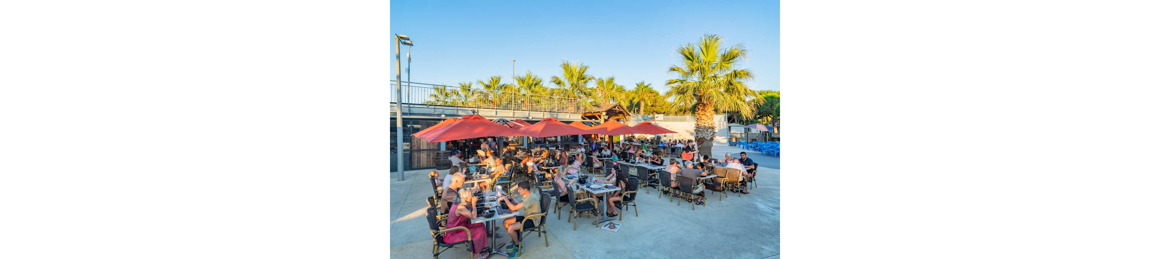 A large group of people dine at an outdoor restaurant terrace under red umbrellas. The setting features palm trees, clear blue skies, and warm evening sunlight. Diners sit at numerous tables enjoying food and drinks in a relaxed atmosphere. The scene conveys a summery, vacation-like mood with a spacious open-air layout.