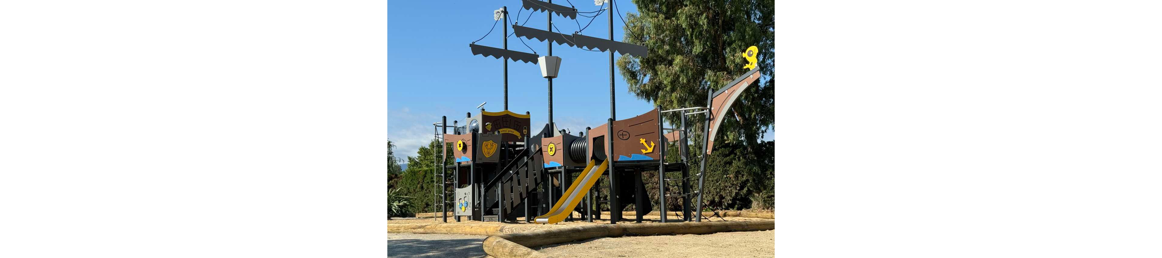 A large pirate ship themed playground structure stands in an outdoor park. The play equipment features multiple decks, slides, and climbing areas styled like a wooden ship. Bright colors such as yellow, blue, and brown highlight anchors, portholes, and playful symbols on the panels. Tall trees and a clear sky frame the background, suggesting a sunny day in a natural setting.