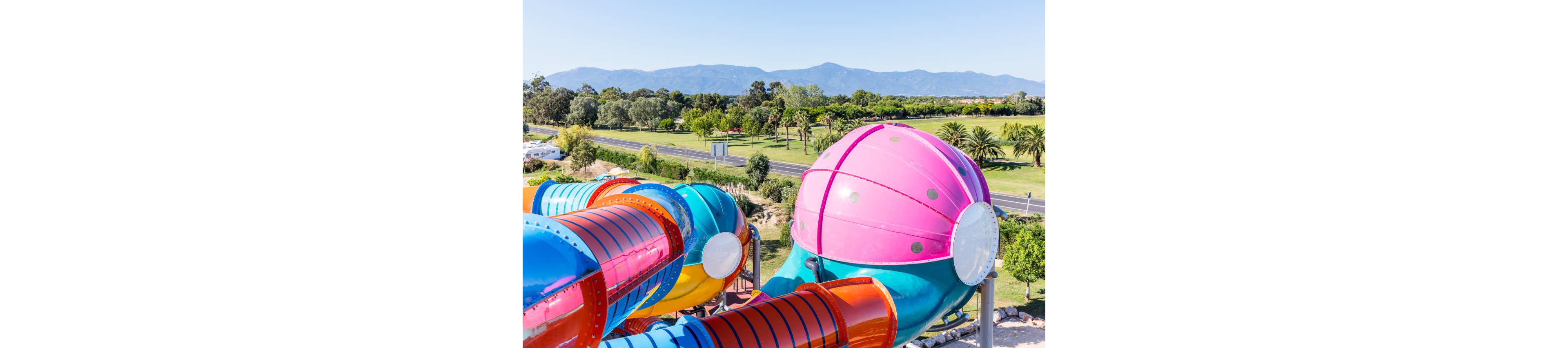 Brightly colored enclosed water slides dominate the foreground of this outdoor scene. The tubes feature vivid pink, blue, orange, and teal sections with a large rounded dome structure at the top. Beyond the slides, a lush green park with trees and palm-lined areas stretches toward a distant road. Mountains rise in the background under a clear blue sky, suggesting a sunny, warm day at a water park.
