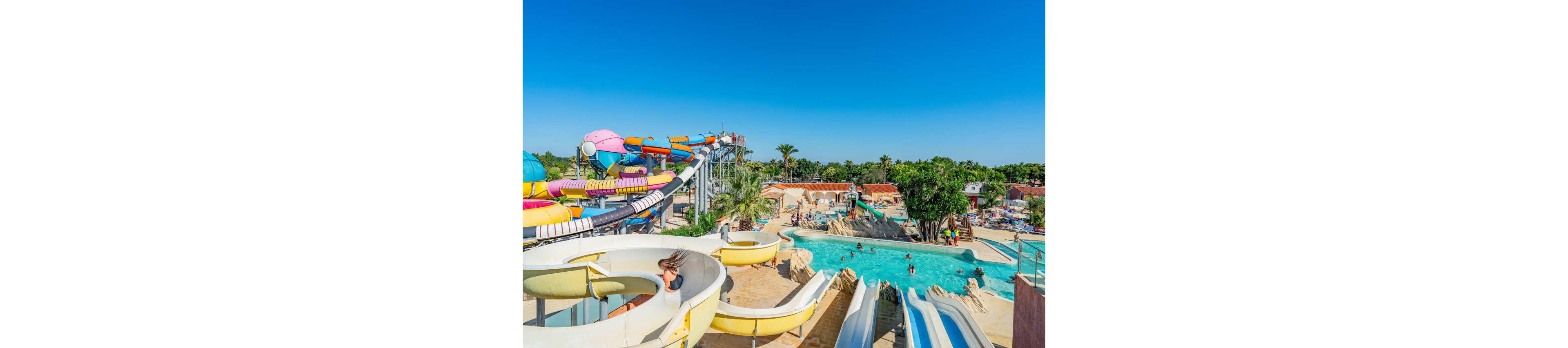 A vibrant outdoor water park features multiple colorful water slides winding above a large pool area. Families and children enjoy swimming, splashing, and sliding under a clear blue sky. Palm trees and tropical landscaping surround the pools, creating a resort-like atmosphere. The bright colors of the slides and sunlit water emphasize a fun, energetic summer setting.
