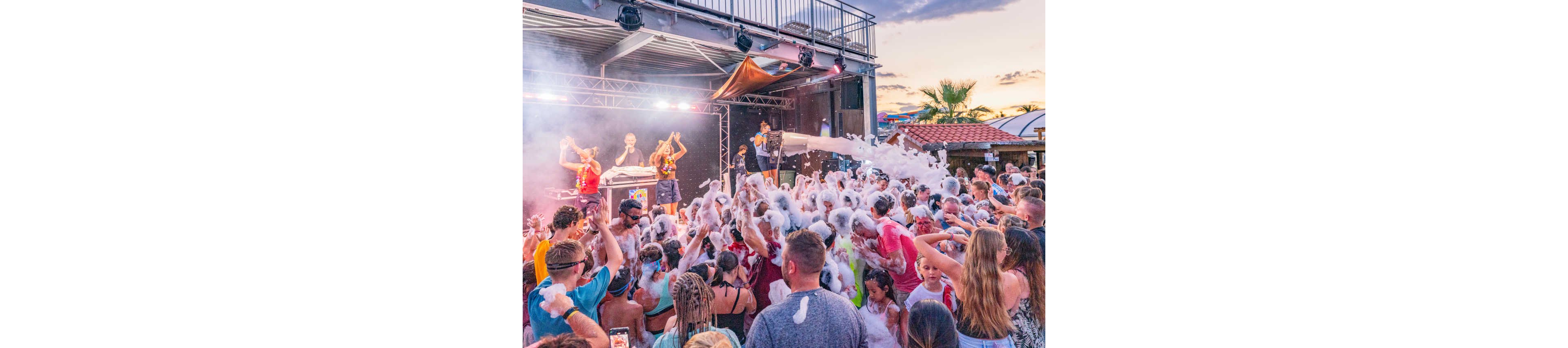 A large crowd of people gathers at an outdoor foam party, covered in thick white bubbles. Performers on a stage spray foam over the audience as they dance and raise their hands. The event takes place at sunset, with colorful stage lights and a partly cloudy sky in the background. The scene feels energetic, festive, and summery, with many families and friends enjoying the lively atmosphere.