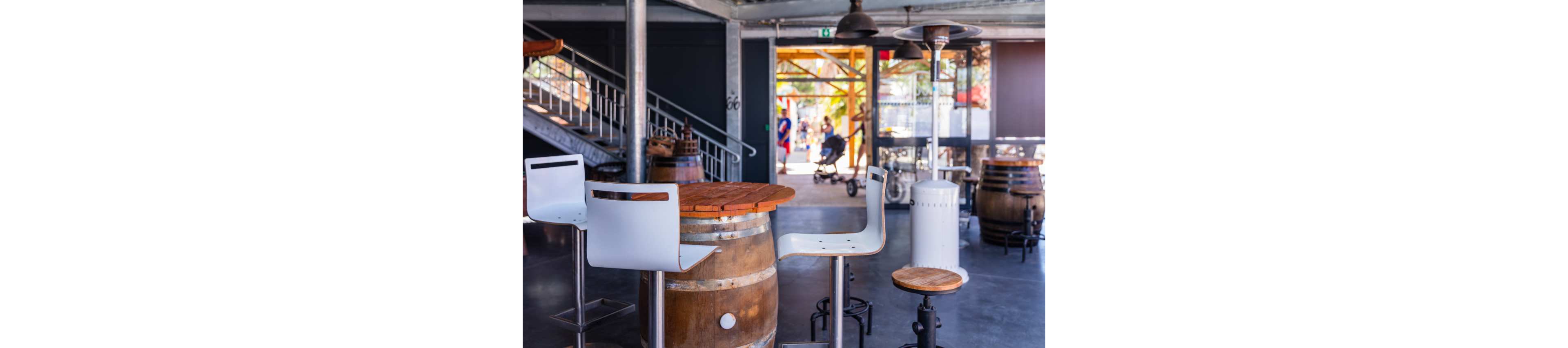 A contemporary bar interior features high stools arranged around a wooden barrel table. Industrial metal beams, concrete flooring, and a staircase create a modern, open atmosphere. Additional barrels and tall heaters are placed throughout the space. Sunlight streams in from the open entrance, where people and a stroller are visible outside, adding a casual, social feel.